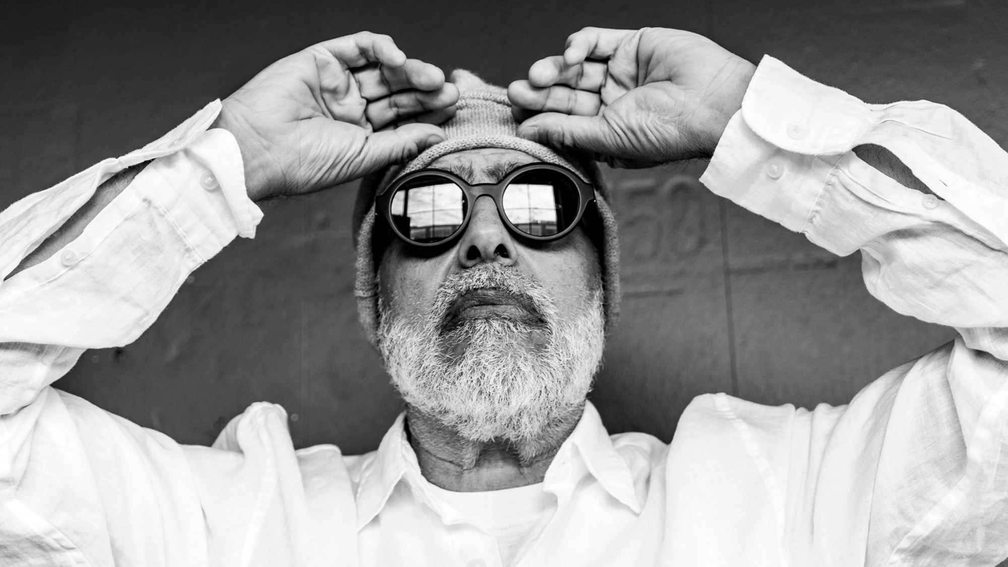 In this high-contrast black and white portrait, theatre composer David Yazbek wears a knit beanie and round sunglasses. He looks upward with his hands to his forehead, dressed in a white button-down shirt against a dark, textured background.