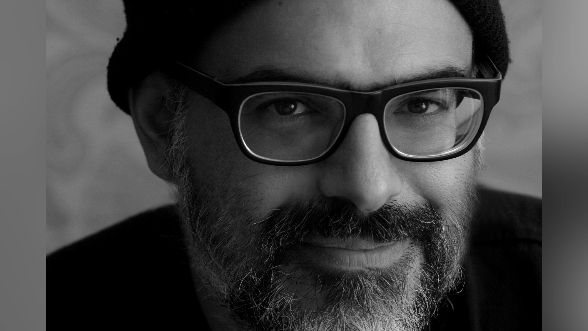 Black-and-white close-up portrait of David Yazbek, a bearded man wearing dark-rimmed glasses and a knit beanie. He is facing the camera and smiling softly. His facial expression appears calm and friendly, and the background is out of focus.