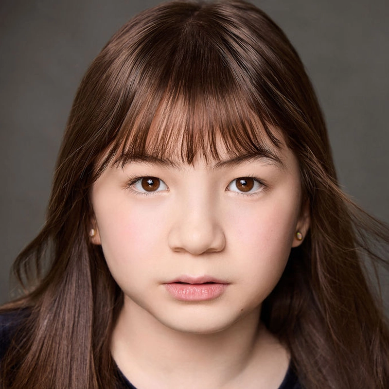 A young girl with straight, brown hair and bangs looks directly at the camera. She has light skin, brown eyes, and a neutral expression. She is wearing small earrings and a dark top. The background is plain and gray. This portrait captures Aerina DeBoer in a calm and contemplative moment.