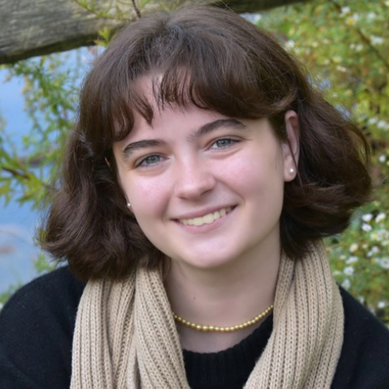 A young person with short brown hair and blue eyes smiles at the camera. They wear a black top, beige scarf, pearl earrings, and a gold necklace. Green foliage and a wooden structure are in the background.