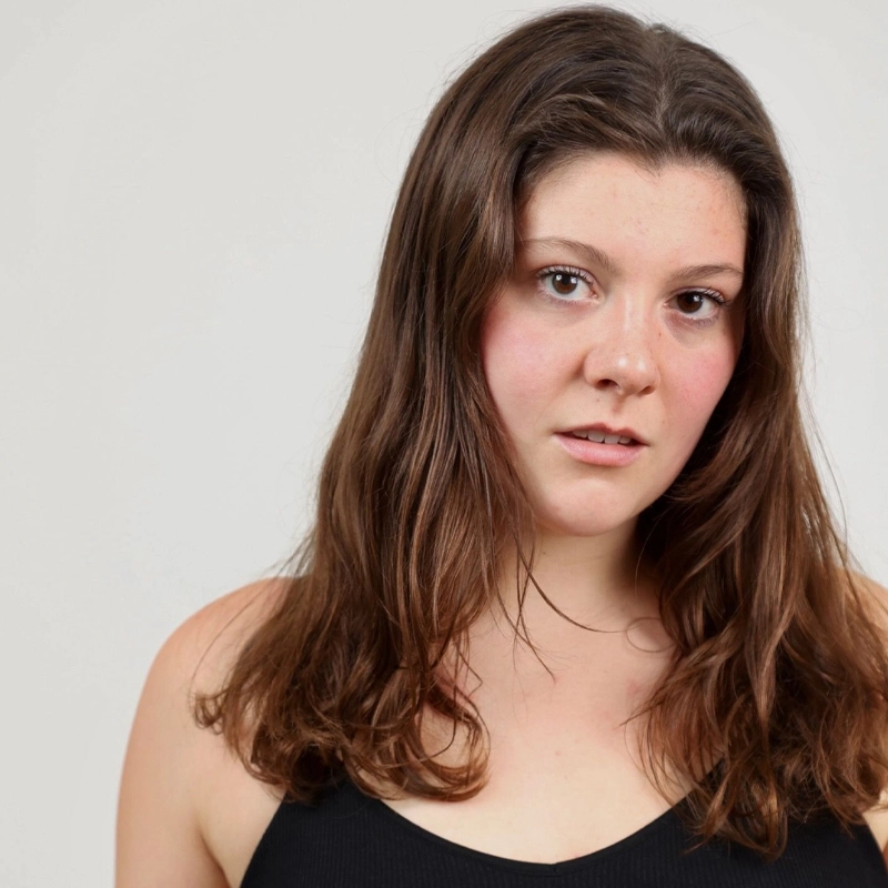 Young woman with long brown hair and fair skin, wearing a black sleeveless top, stands against a plain light gray background. Katie Dinsmore looks at the camera with a neutral expression and slightly parted lips. Her cheeks have a natural, rosy tint.