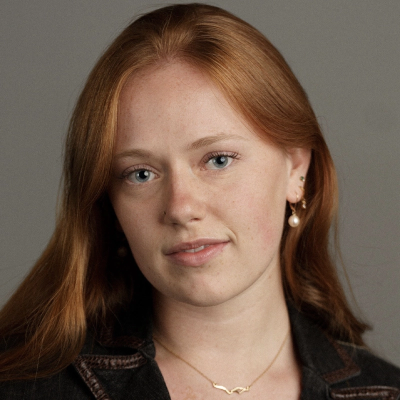 A young woman with long red hair, light skin, and blue eyes looks into the camera. She wears gold hoop and pearl earrings, a gold necklace, and a dark jacket. The background is plain gray. Her expression is calm and neutral.