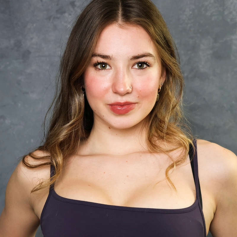 Sloane Escobar, a young woman with long brown hair and light skin, stands against a gray textured background. She has a small nose ring, subtle makeup, and wears a dark tank top. Sloane looks directly at the camera, smiling softly.