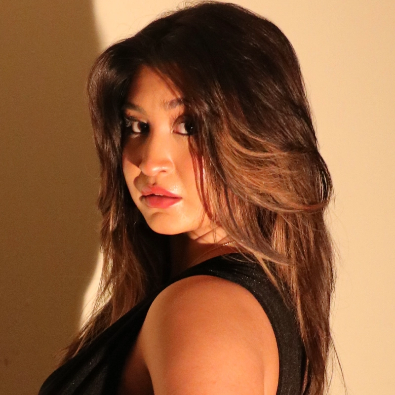 Ava Luisa Estrada, a woman with long brown hair and highlighted strands, looks over her shoulder toward the camera, lips parted. She wears a sleeveless black top and stands against a beige wall with dramatic lighting creating shadows on the background.
