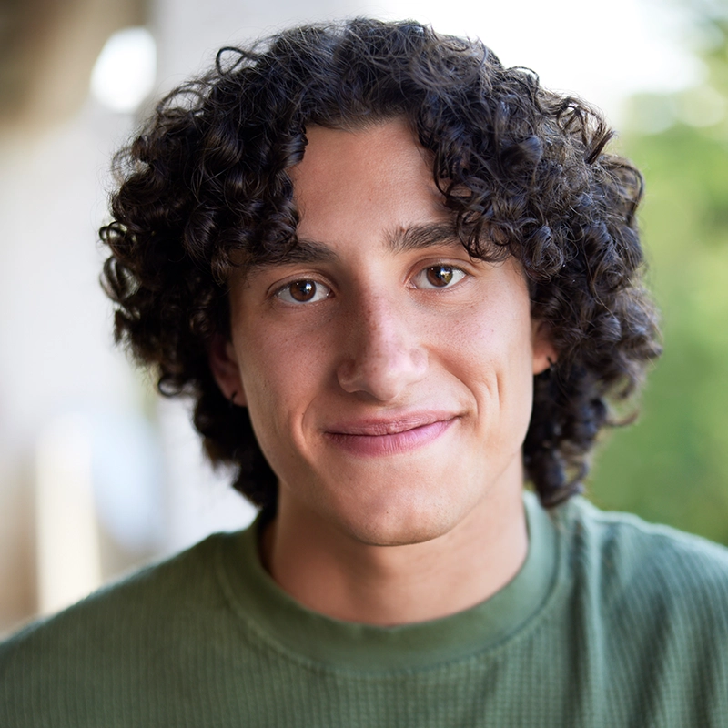 Young adult Celeste Zwingel, with curly brown hair, wears a light green textured shirt and smiles softly at the camera outdoors. The background is blurred with greenery and light, highlighting Celeste’s relaxed and friendly expression.