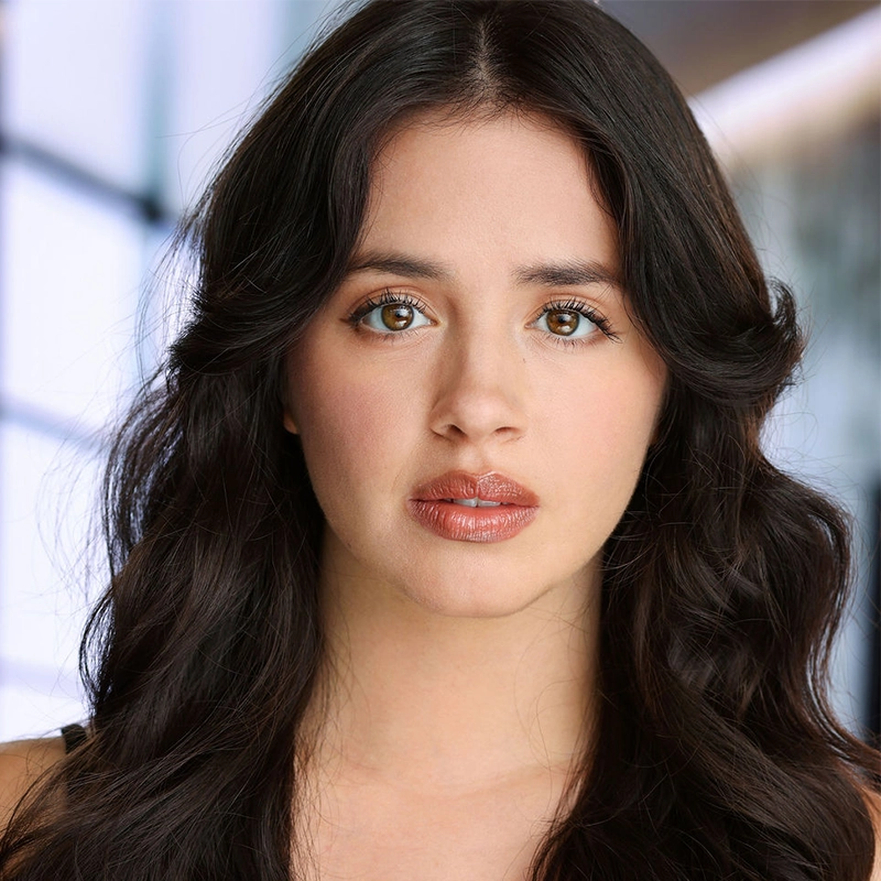 A young woman with long, wavy dark hair and light brown eyes looks directly at the camera. She has natural makeup, defined brows, and glossy lips. The background is blurred with soft lighting and geometric window shapes, creating a modern, airy feel—capturing the poise and presence reminiscent of Fabi Aguirre.