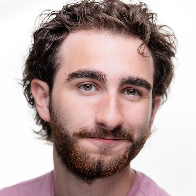 Young man with wavy brown hair and a full beard smiles softly at the camera. He has thick eyebrows, light brown eyes, and wears a light pink shirt. The background is bright white, making his features stand out clearly.