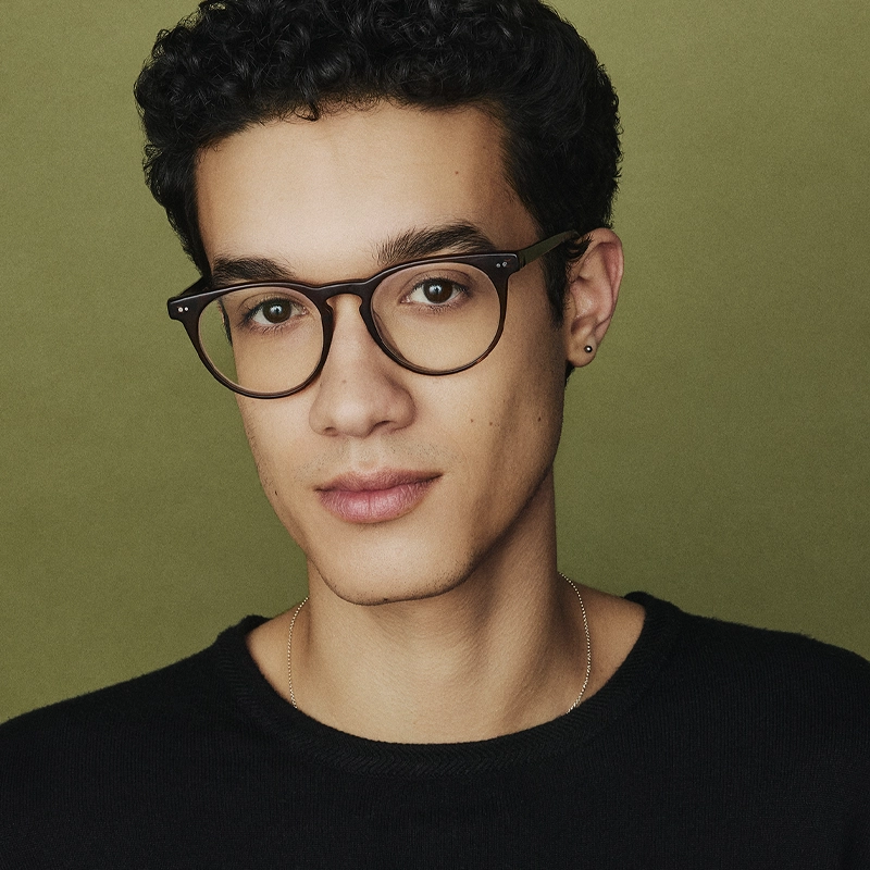 A young person with short curly dark hair, wearing round glasses, a small earring, and a thin necklace, gazes softly at the camera. They are dressed in a black top, with a neutral olive green background behind them.