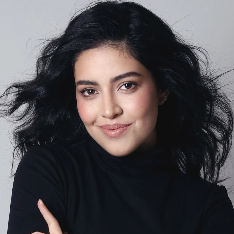 A woman with wavy black hair and fair skin wears a black turtleneck. She smiles gently, looking at the camera. Her makeup is natural with rosy cheeks and nude lips, capturing Elena Reyes Farias’s understated elegance. The background is a neutral light gray, and her hair fans out softly around her head.