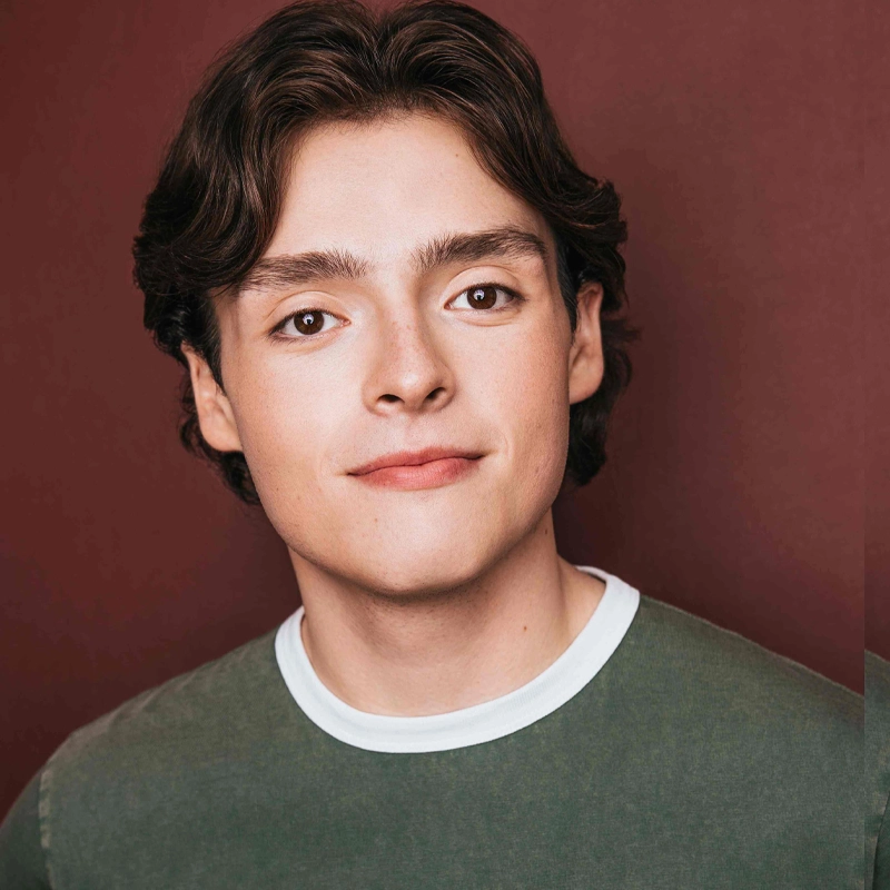 A young man with wavy brown hair smiles softly at the camera. He wears a green crewneck shirt with white trim. The background is a solid reddish-brown color. His expression is relaxed and friendly, capturing the approachable demeanor of Jackson Flaherty.