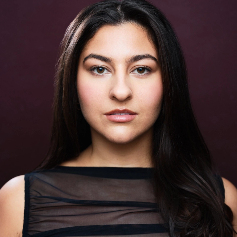 Nicole Fluegge, a woman with long dark hair and fair skin, looks directly at the camera. She wears a sheer black top with horizontal stripes. The background is a deep maroon color. Her expression is neutral and her makeup is natural.