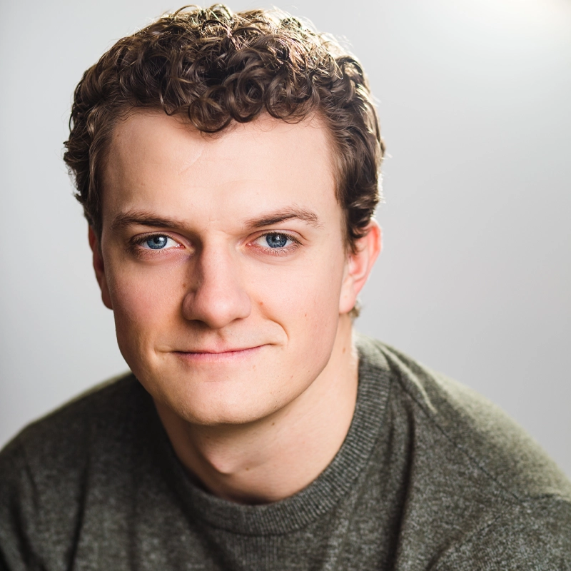 A person with short, curly brown hair and blue eyes smiles softly at the camera. They are wearing a green sweater and are posed in front of a plain, light gray background with soft lighting.