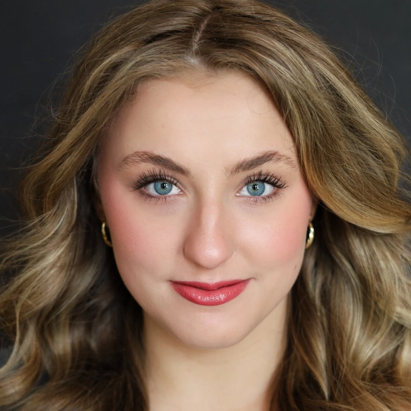 Ella Grace Francis, a young woman with wavy, light brown hair and blue eyes, smiles softly. She wears gold hoop earrings, pink blush, and glossy lipstick. Her makeup is subtle and natural. The background is dark and plain, highlighting her facial features.