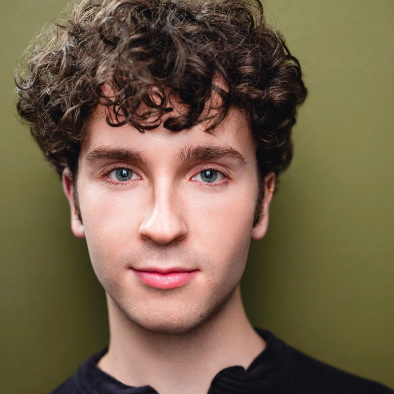A young person with fair skin, blue eyes, and curly brown hair is shown facing forward. They are wearing a black top and smiling slightly. The background is a muted olive green color. The lighting is soft and even. 297 characters.