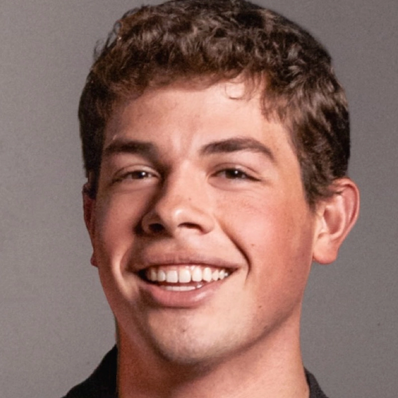A young man with short, curly brown hair smiles warmly at the camera. He has fair skin, brown eyes, and is wearing a dark shirt. The background is plain gray, and the lighting is even, highlighting his friendly expression.