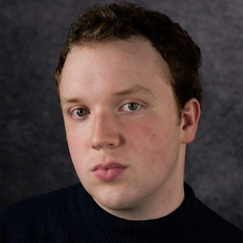 Young adult with fair skin, short curly brown hair, and hazel eyes looks directly at the camera with a neutral expression. Wearing a dark sweater, they are posed against a dark gray, textured background. Lighting highlights the face evenly.