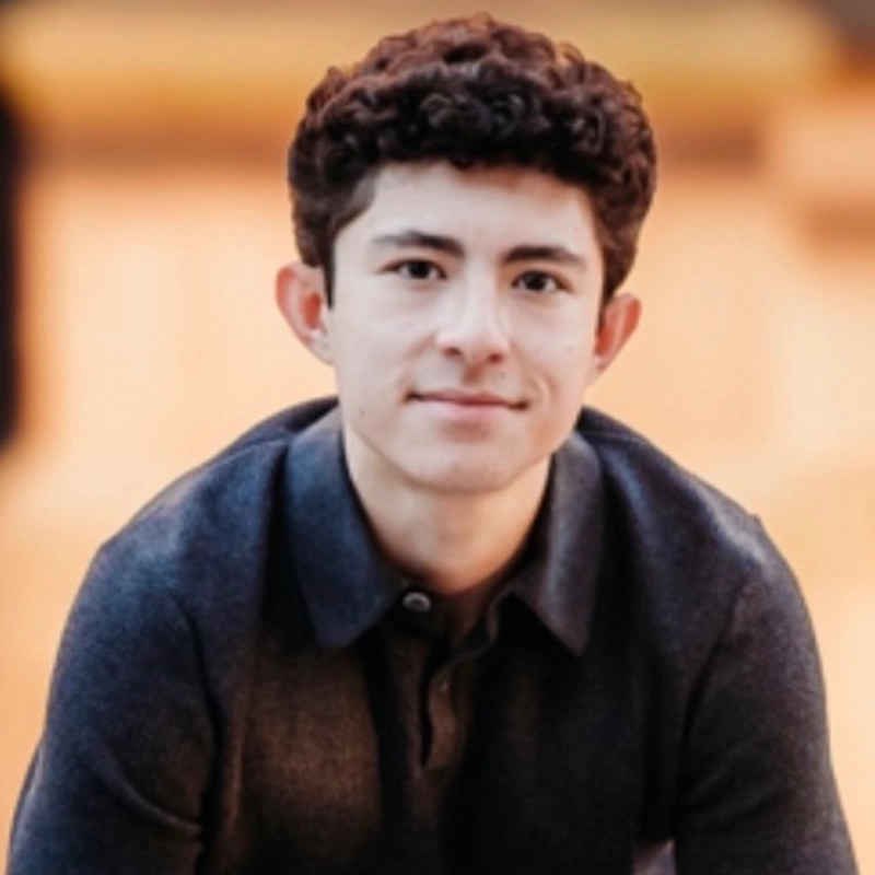 Young man with short, curly brown hair and fair skin, wearing a dark collared shirt, looks at the camera and smiles softly. The background is blurred in shades of beige and orange, drawing focus to his face and upper body—a portrait reminiscent of Tyler Joseph’s understated style.