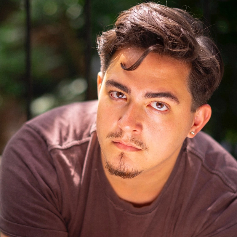 A young man with short brown hair, light skin, and facial hair looks directly at the camera. He wears a brown T-shirt and a small hoop earring. The background is softly blurred greenery, suggesting an outdoor setting on a sunny day.