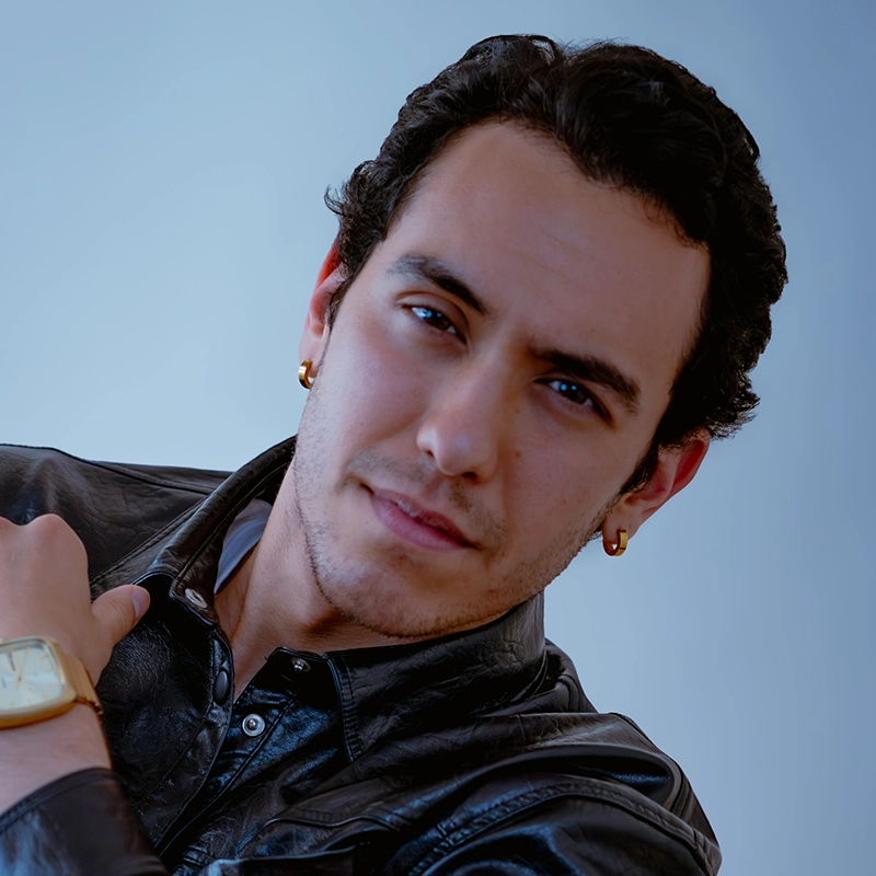 Salvador Diaz-DeBose, a man with short dark curly hair, light skin, and gold hoop earrings, wears a black leather jacket and a gold watch. He is looking directly at the camera with a slight, confident smile against a plain light blue background.