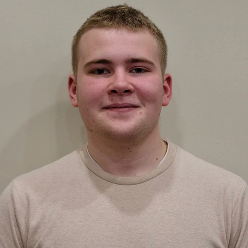 A young person with short light brown hair and fair skin smiles slightly while facing the camera. Reminiscent of Joseph Gillhooly, they are wearing a beige t-shirt and standing against a plain, light-colored wall. The lighting is even and soft.