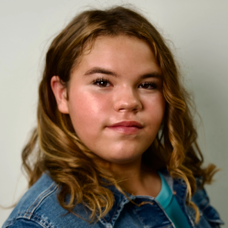 A young person with wavy, light brown hair poses against a plain background. They wear a blue denim jacket over a turquoise top and have a neutral, confident expression reminiscent of Kelsey Valente. Their head is tilted slightly to the right, and their hair falls over their shoulders.