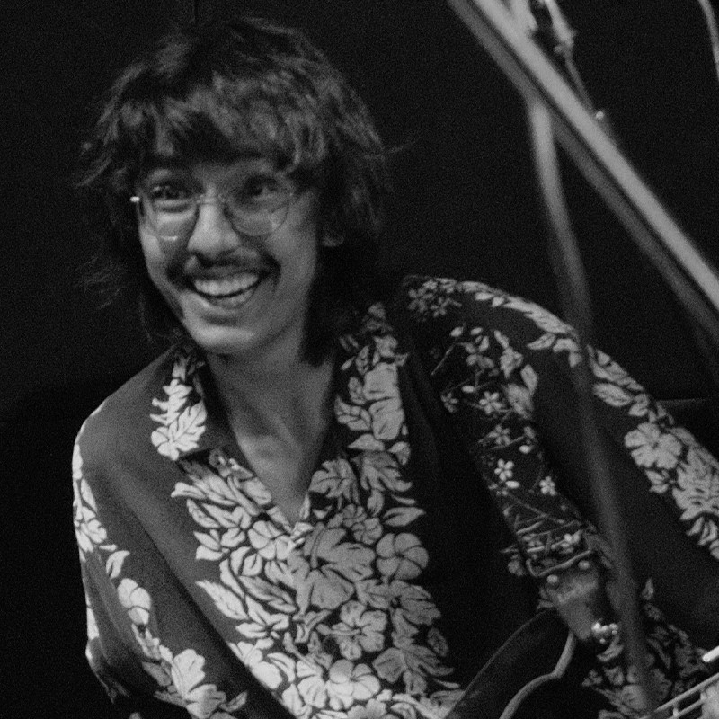 Black and white photo of Kenji Golden, a smiling person with wavy hair, glasses, and a mustache. They wear a floral-patterned shirt and appear to be holding a guitar. Microphone stands or cables are visible in the foreground.