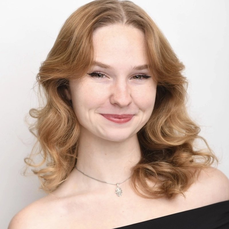 Tori Greenlaw, a young woman with wavy, shoulder-length light brown hair, smiles softly. She wears a black off-the-shoulder top and a silver necklace with a Hamsa hand charm. The background is plain white, and her expression is warm and friendly.