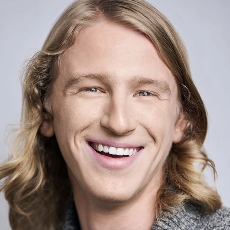 A smiling person with long, wavy blonde hair, fair skin, and blue eyes is wearing a gray top. The background is a soft, light gray, and the lighting is bright, giving the portrait a cheerful and friendly atmosphere—perfectly capturing the warm personality of Dylan Grosh-Hoy.