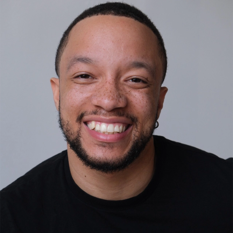 A smiling man, Izaiah Harris, with short black hair, a trimmed beard, and freckles wears a black shirt. He faces the camera against a plain, light gray background. The lighting is even, and he appears happy and approachable.