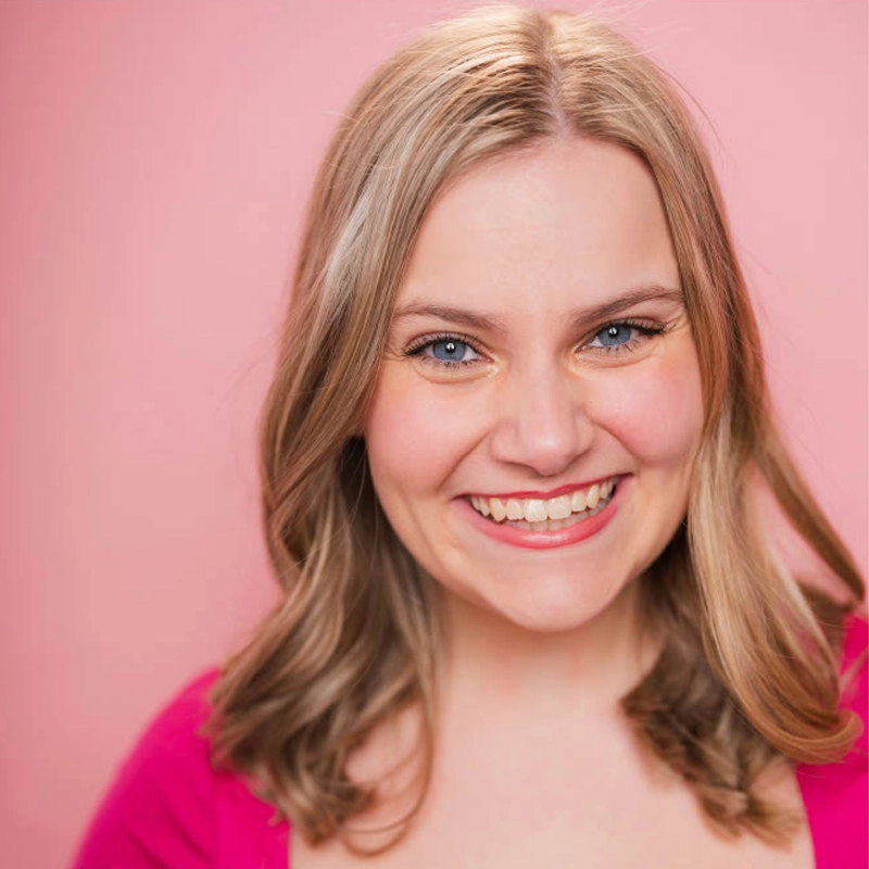 A smiling woman with shoulder-length blonde hair stands against a solid pink background. She wears a bright pink top and has blue eyes, light makeup, and a cheerful expression. Her hair is parted slightly to the side and styled in loose waves.