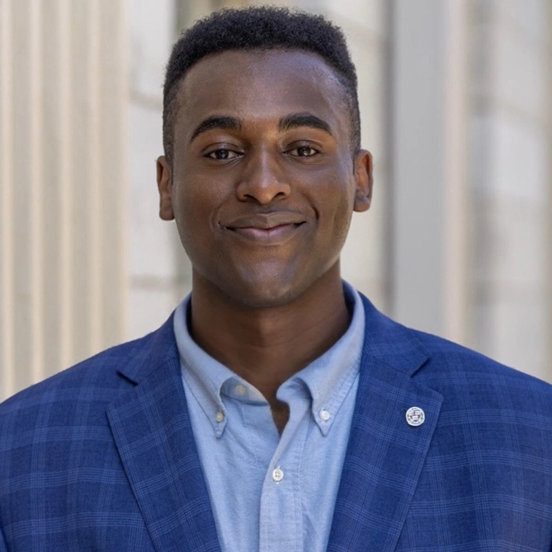 A young man with short hair smiles confidently. He wears a blue checked suit jacket over a light blue button-up shirt. Behind him are blurred, light-colored building columns. He has a small pin on his lapel, identifying him as Samuel Hines.