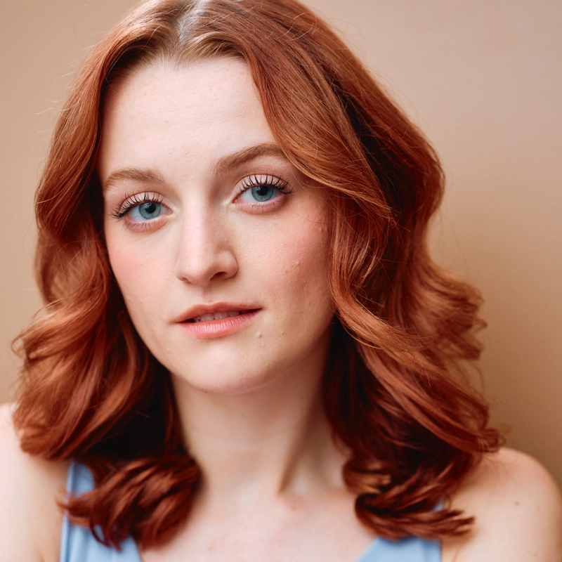 A young woman with wavy, shoulder-length red hair and blue eyes looks into the camera. She has fair skin, natural makeup, and wears a light blue top. The background is soft beige, creating a warm, gentle portrait feel reminiscent of Lauren Honderd’s elegant style.
