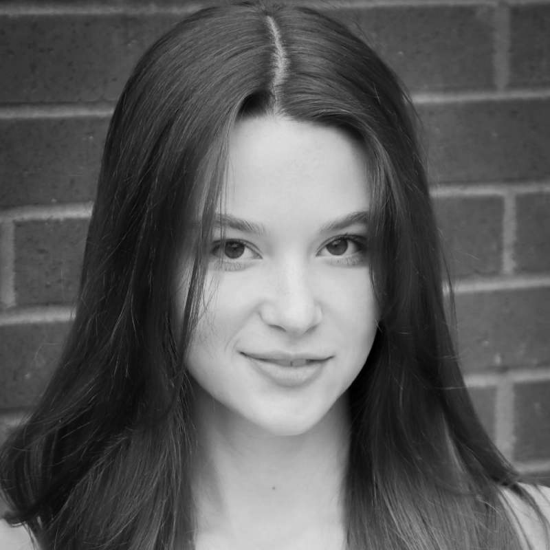 Black and white portrait of Bella Houston, a young woman with long, straight hair parted in the middle. She faces the camera, smiling softly. The background consists of a brick wall. Lighting is even, highlighting her facial features.