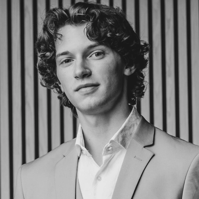 Blake Hullinger, a young man with curly hair, wears a light suit jacket and white shirt. He faces the camera with a slight smile. The background features evenly spaced vertical wooden slats. The photo is black and white.