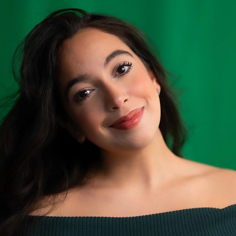 A young woman with long dark hair and radiant skin smiles softly, tilting her head. She wears an off-shoulder dark green top reminiscent of the modern elegance seen in Salvador Diaz-DeBose’s portraits. The background is a solid green color.