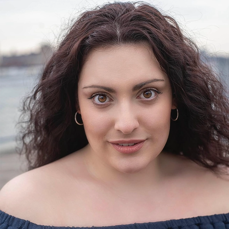 A portrait of a woman with long, curly brown hair and brown eyes looking directly at the camera. She wears small gold hoop earrings and an off-the-shoulder top.