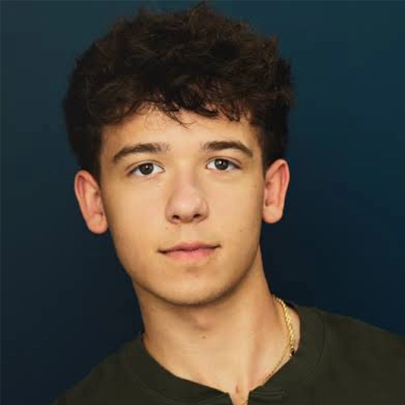 A young person with short, curly dark hair and light skin gazes at the camera with a neutral expression. They wear a dark shirt and a thin gold necklace. The background is a smooth, dark blue gradient.