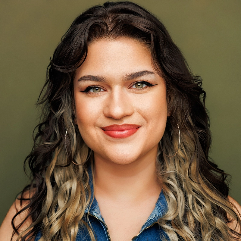 Smiling person with long wavy brown hair featuring lighter tips, wearing large hoop earrings and a sleeveless denim top, poses against an olive green background. The person has winged eyeliner, defined brows, and natural makeup with red lipstick. 300 characters.