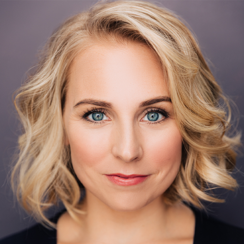 A woman with short, wavy blonde hair and blue eyes looks directly at the camera, smiling softly. She wears natural makeup and a dark top. The background is plain and softly lit, keeping the focus on her face.
