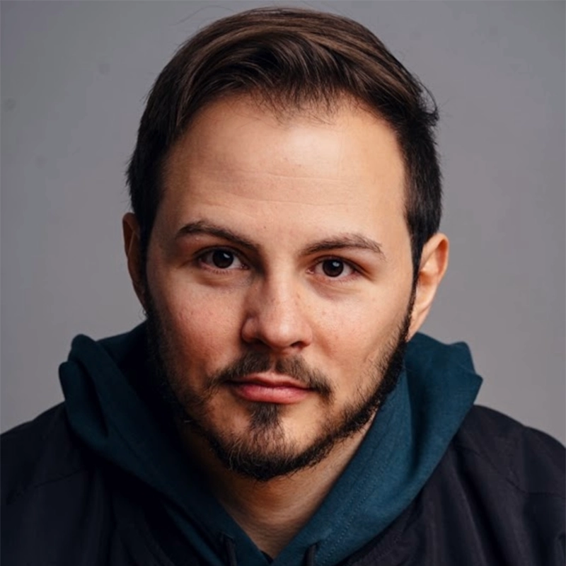 Kendall Tubbs, a man with short brown hair, neatly trimmed beard, and mustache, stares directly at the camera. He wears a dark jacket over a teal hoodie. The background is plain and gray, and the lighting highlights his facial features clearly.