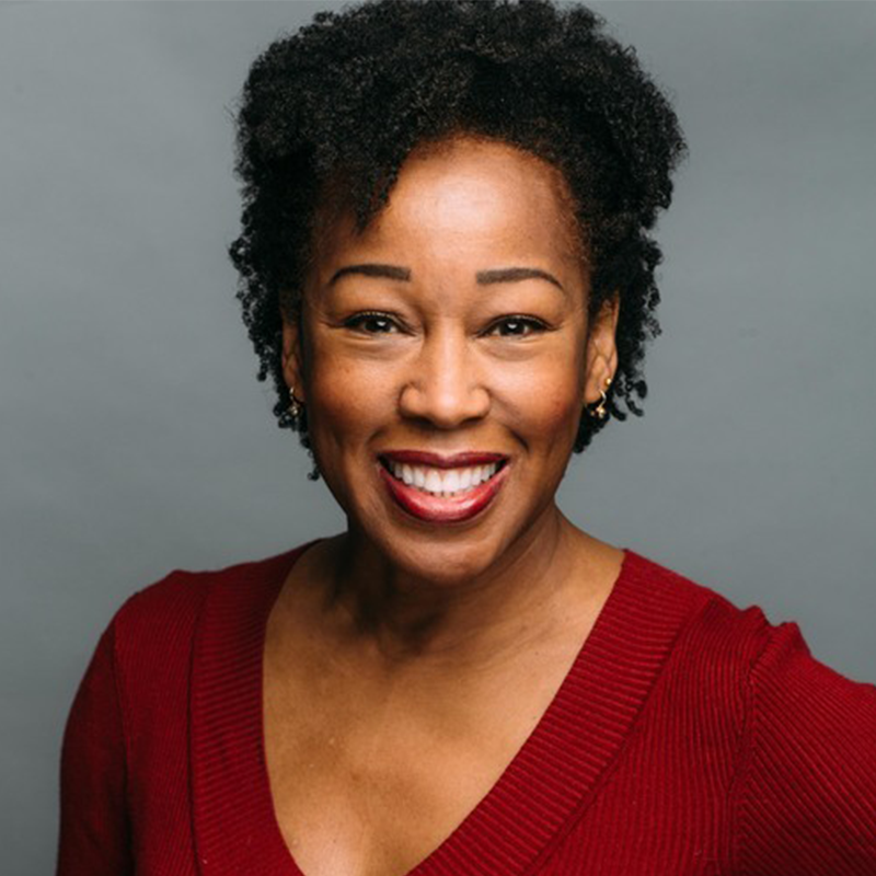 A woman with short curly hair smiles warmly. She wears a deep red V-neck sweater and small earrings. The background is solid gray, and she is looking directly at the camera, conveying confidence and friendliness.