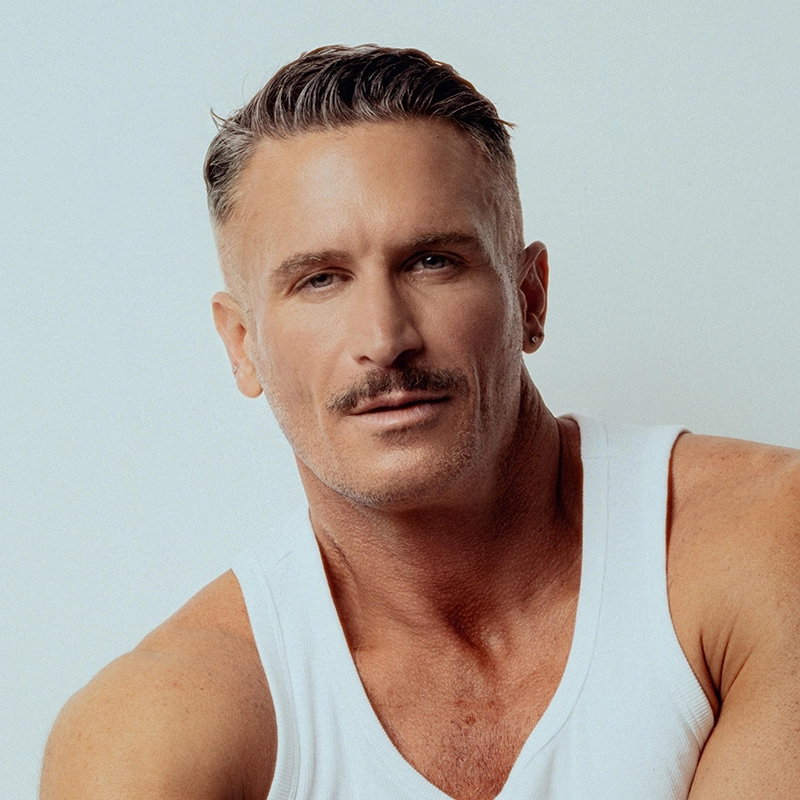 A middle-aged man with short, styled brown hair and a mustache wears a white tank top. He has light skin, blue eyes, and a small earring in his left ear. John Arthur Hill looks confidently at the camera with a neutral background behind him.