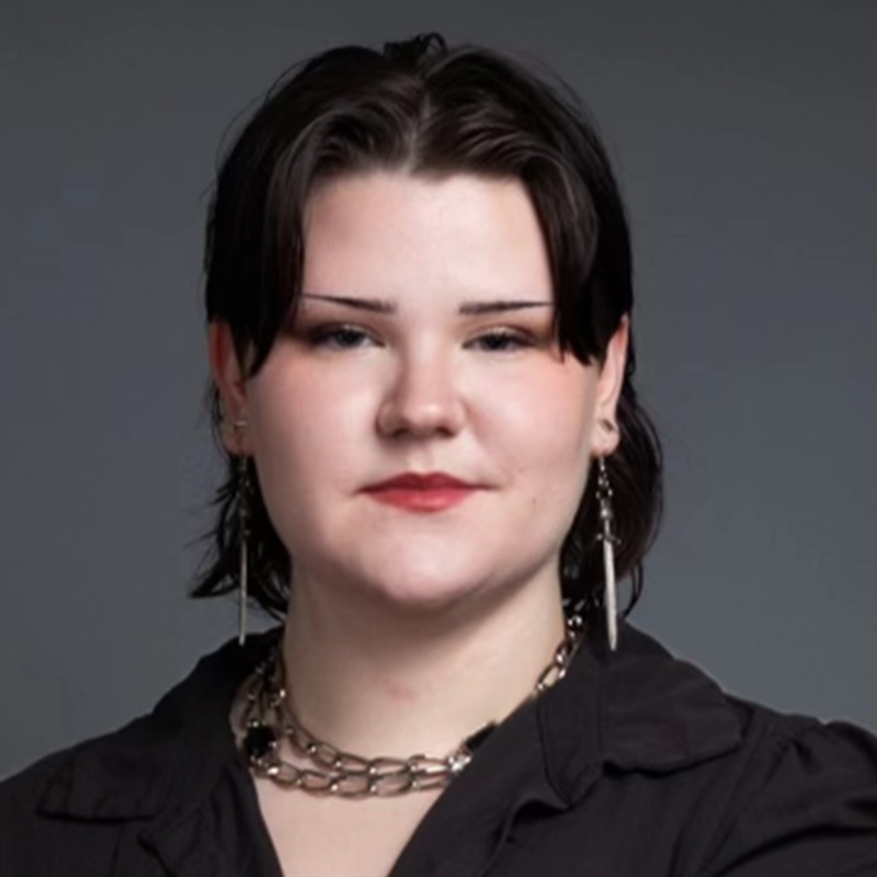 A person with dark hair and thin eyebrows faces the camera. They wear a layered chain necklace and earrings shaped like small swords.