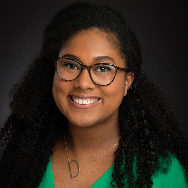 A woman with long curly hair, wearing glasses and a green top, smiles at the camera. She has a gold necklace with a large “D” pendant and small earrings. The background is dark, highlighting her face and features—capturing the warmth and confidence of Danielle Victoria Kaigler.