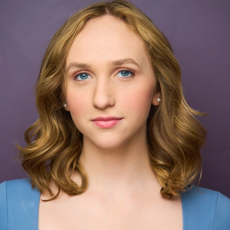 A woman with wavy, shoulder-length blonde hair and blue eyes looks at the camera against a purple background. She wears a blue top, pearl earrings, and has a calm, neutral expression. Her makeup is natural, with pink lipstick and subtle eye makeup. This portrait features Katie Kallay.