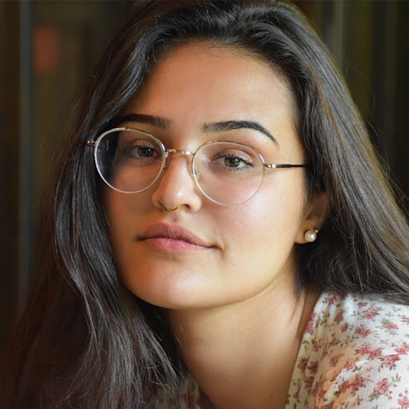 A young woman with long dark hair wears thin-rimmed glasses, a small gold septum ring, and pearl earrings. She is looking directly at the camera.