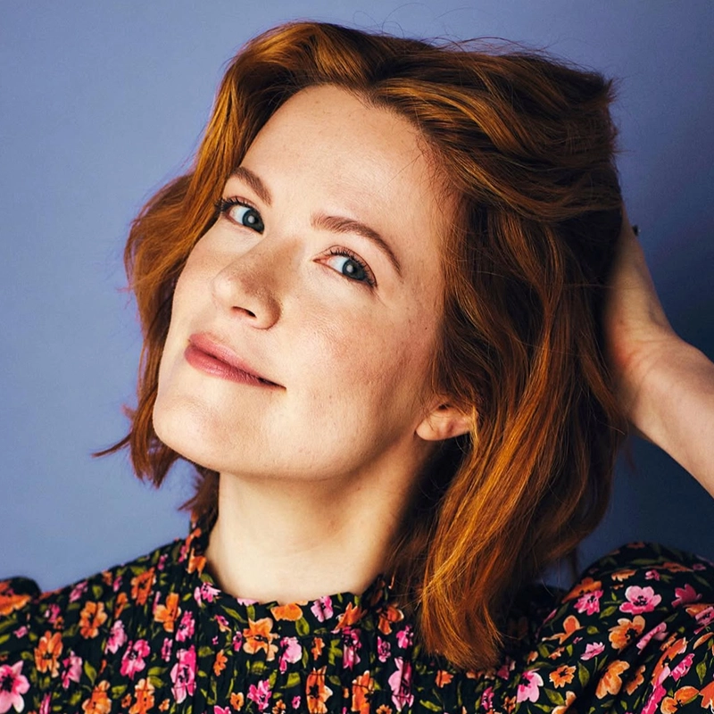 A woman with shoulder-length auburn hair looks at the camera with a gentle smile. She has one hand raised to her hair and wears a black blouse with a colorful floral print.