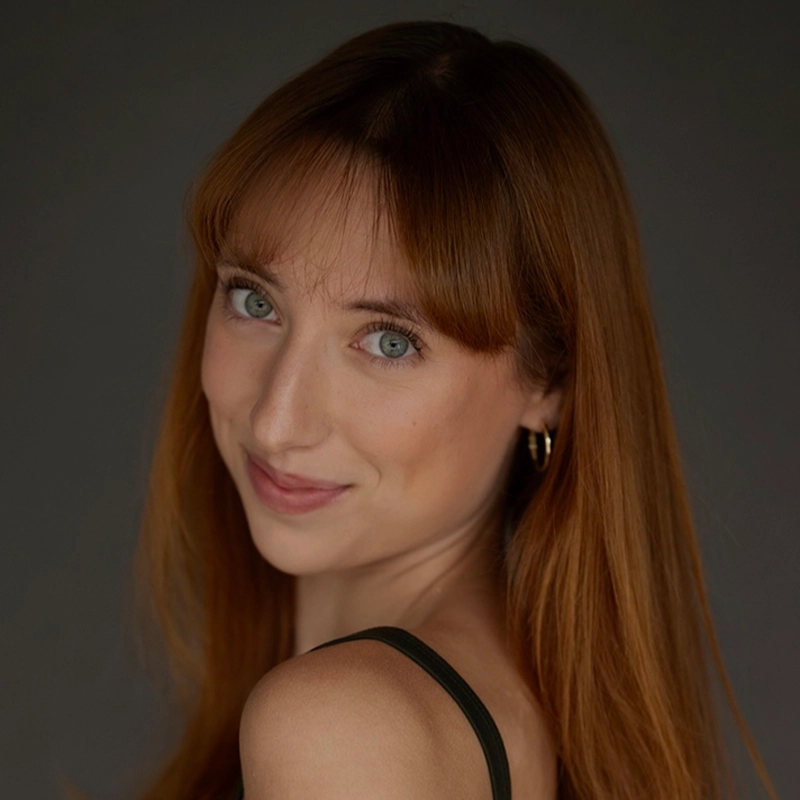 A young woman with long straight auburn hair, fair skin, and light green eyes smiles softly at the camera. She wears a black sleeveless top and gold hoop earrings, with a neutral gray background behind her.
