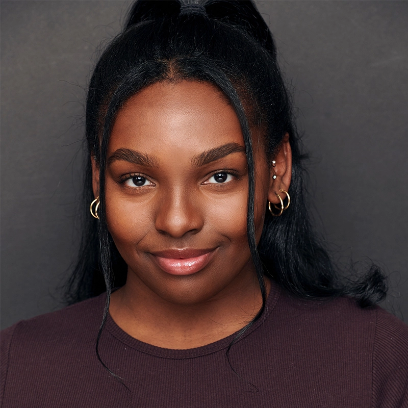 Alyssa Enita Stanford, a young woman with dark skin, smiles gently. Her long black hair is tied back, accentuating her smooth skin. She wears small gold hoop earrings and an ear cuff, paired with a dark top against a plain dark background.