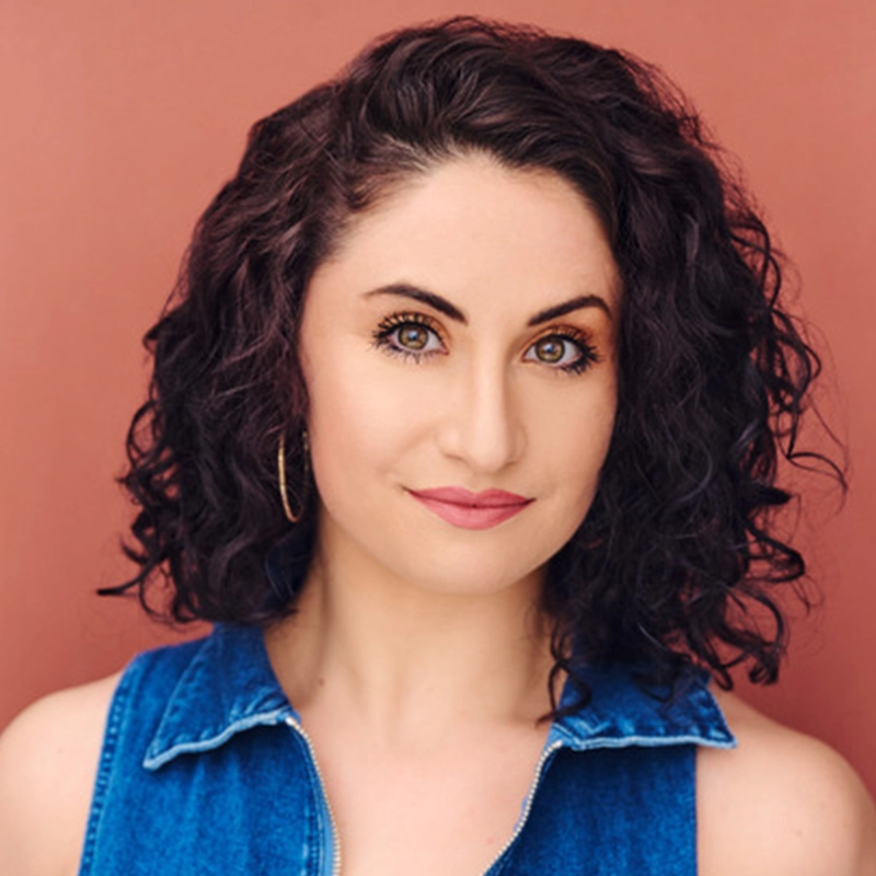 A woman with curly, dark brown hair and fair skin smiles slightly at the camera. Kelly MacMillan wears a sleeveless blue denim top and gold hoop earrings. The background is a solid, muted pink color.